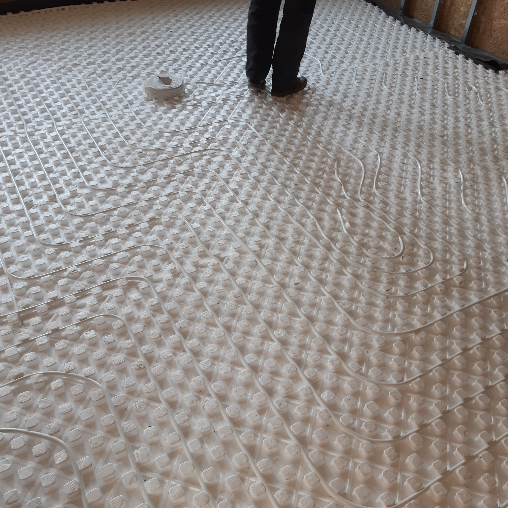 Biała mata izolacyjna z ułożonymi rurami ogrzewania podłogowego, widoczne nogi instalatora w czarnych spodniach i butach roboczych oraz fragment ściany w tle.