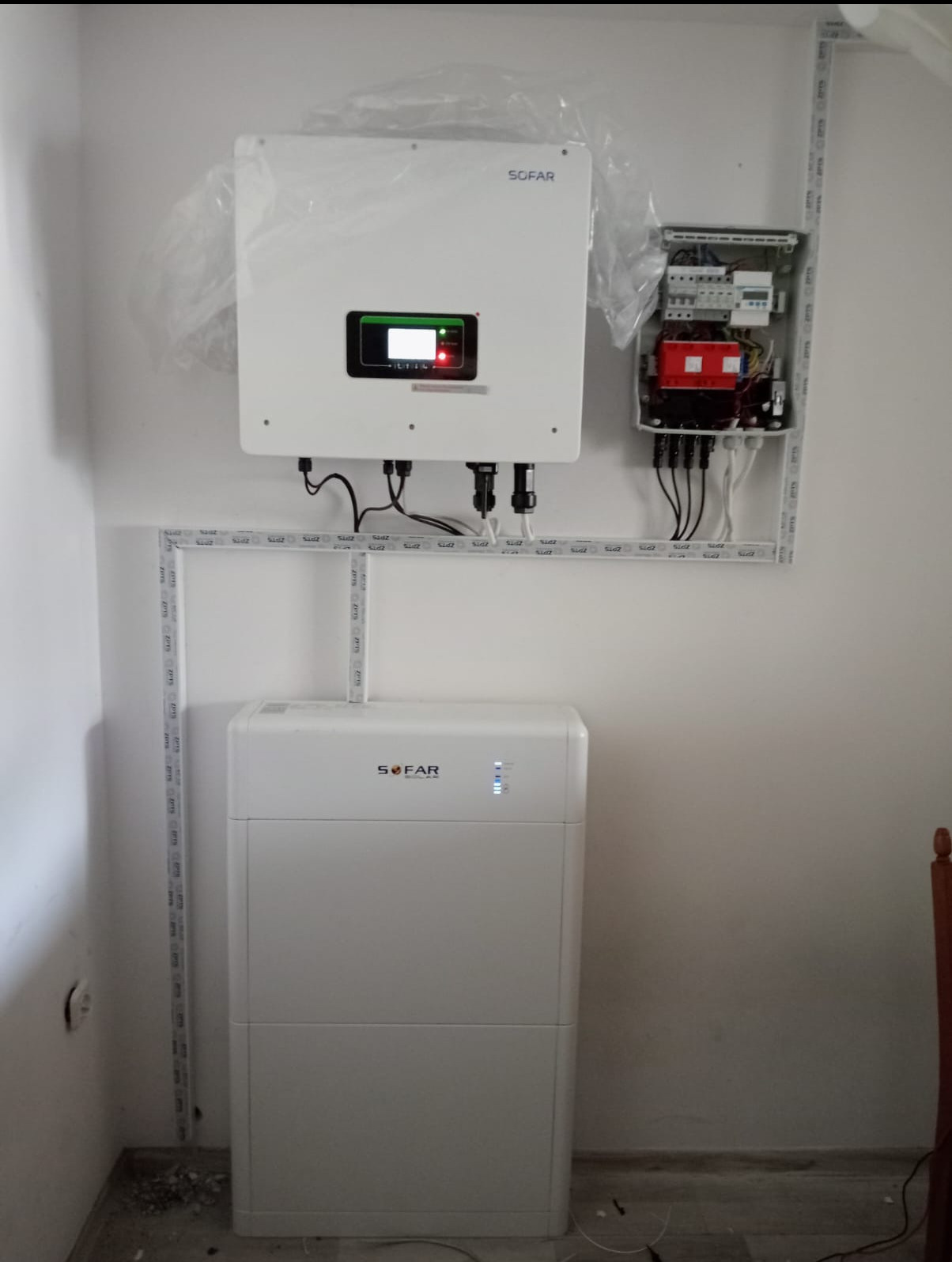 Biała instalacja z falownikiem i akumulatorem marki Sofar, wraz z rozdzielnicą elektryczną, zamontowana na ścianie wewnątrz budynku. Instalacja zabezpieczona folią.