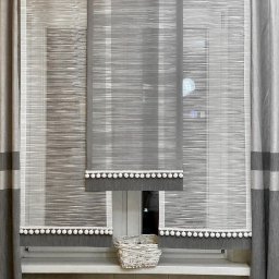 Dom i k sp. z oo - Szare, poziomo tkane rolety rzymskie z białymi pomponami, uzupełnione zasłonami w odcieniach szarości z poziomymi pasami, na tle okna z koszykiem na parapecie.