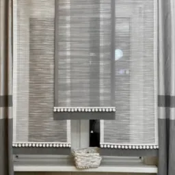 Szare, poziomo tkane rolety rzymskie z białymi pomponami, uzupełnione zasłonami w odcieniach szarości z poziomymi pasami, na tle okna z koszykiem na parapecie.