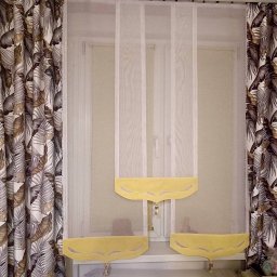 Dom i k sp. z oo - Okno z białymi, transparentnymi zasłonami z żółtymi falbanami i zasłonami po bokach w motywie liści palmowych.