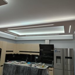 Dom i k sp. z oo - Remontowana kuchnia z osłoniętym blatem, widoczne narzędzia i akcesoria malarskie, nowoczesne oświetlenie sufitowe, zabudowa kuchenna w jasnych kolorach i srebrna lodówka.