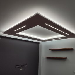 Dom i k sp. z oo - Kraków: Nowoczesny sufit podwieszany z oświetleniem LED w kształcie kwadratu, brązowe elementy dekoracyjne, minimalistyczne półki na ścianie, czarna ściana w tle.