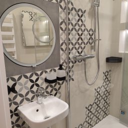 Dom i k sp. z oo - Nowoczesna łazienka z okrągłym lustrem, umywalką nablatową, kabiną prysznicową i płytkami w geometryczny wzór.
