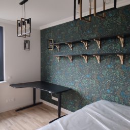 Dom i k sp. z oo - Nowoczesny pokój z czarnym biurkiem z regulowaną wysokością, półkami na tle wzorzystej tapety i designerskimi lampami wiszącymi; meble częściowo zakryte folią ochronną.