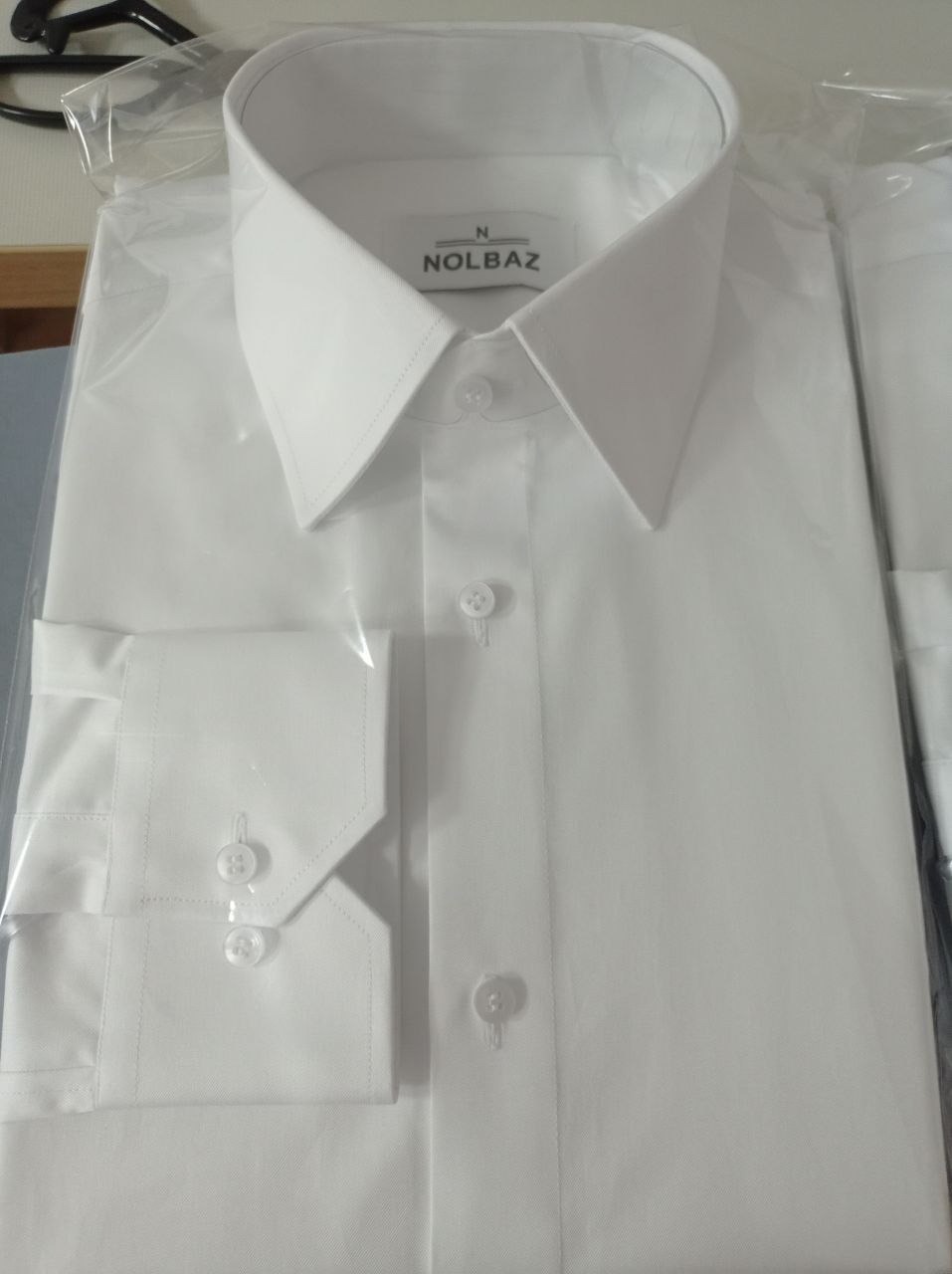 Biała, nowa koszula męska marki NOLBAZ, zapakowana w folię ochronną, widoczne mankiety z guzikami.