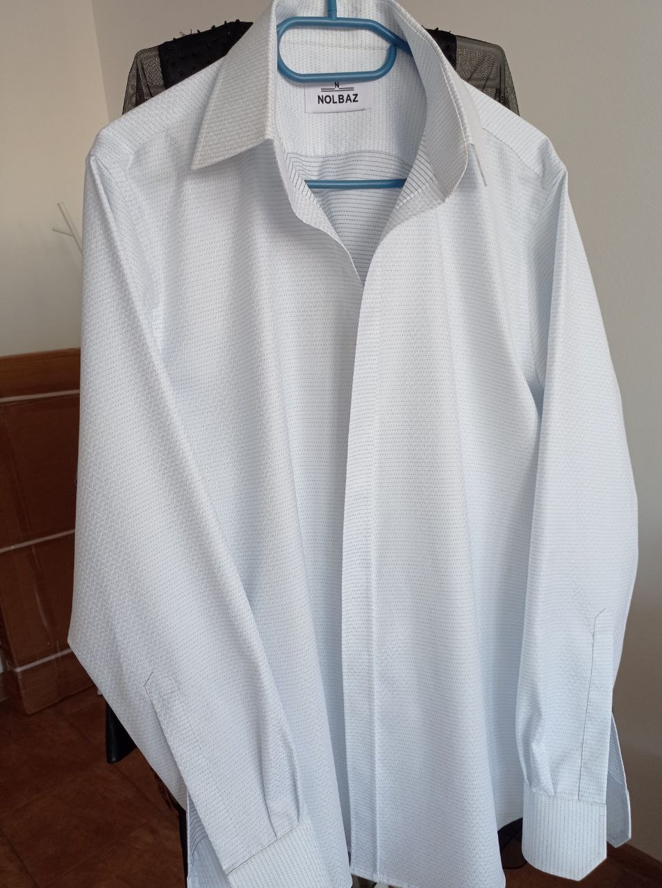 Biała koszula męska z długim rękawem, wisząca na niebieskim wieszaku z logo 'NOLBAZ' widocznym na metce kołnierzyka.