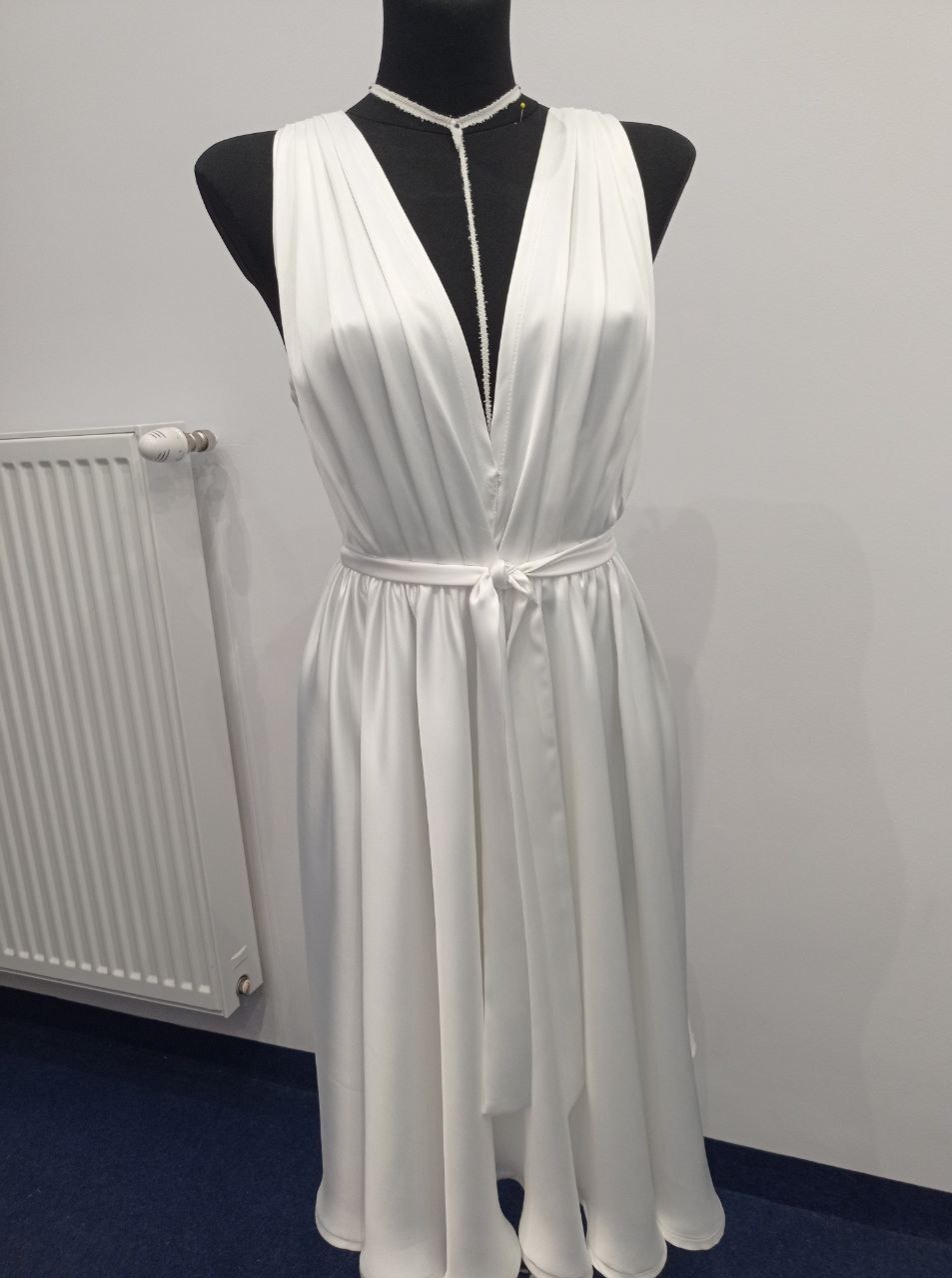 Elegancka, długa suknia ślubna z głębokim dekoltem w kształcie litery V, uszyta z białego, lejącego się materiału, zaprezentowana na czarnym manekinie krawieckim, z widocznym grzejnikiem w tle.