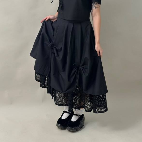 Czarna spódnica z bawełny z koronkowym wykończeniem, zdobiona kokardami, prezentowana na modelce w białych skarpetkach i czarnych butach na platformie.