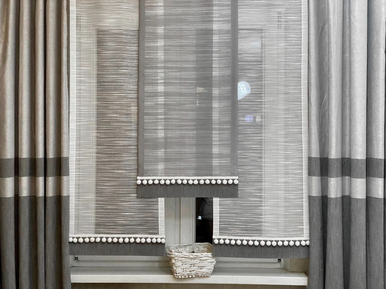 Szare, poziomo tkane rolety rzymskie z białymi pomponami, uzupełnione zasłonami w odcieniach szarości z poziomymi pasami, na tle okna z koszykiem na parapecie.