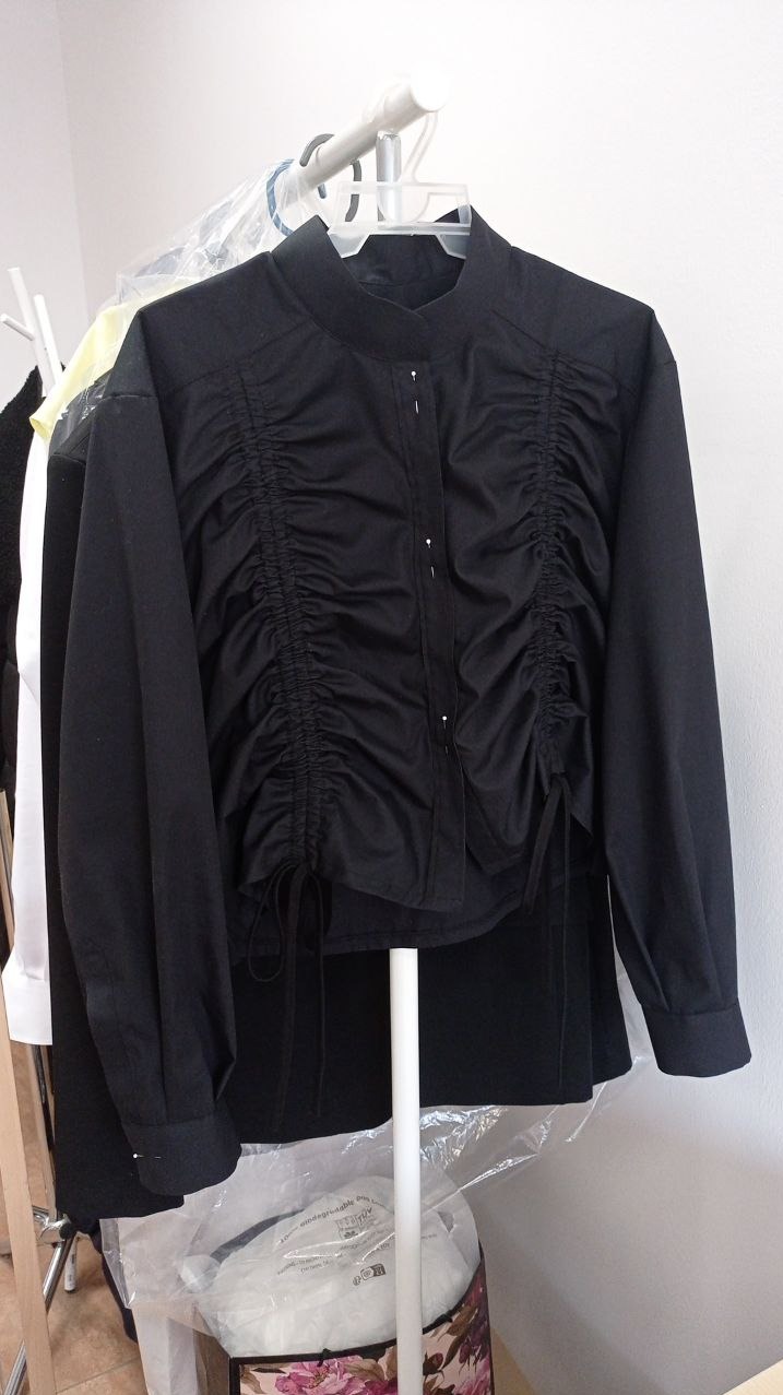 Czarna bluzka z marszczeniami i wiązaniami na wieszaku w szwalni, widoczne inne ubrania w tle.