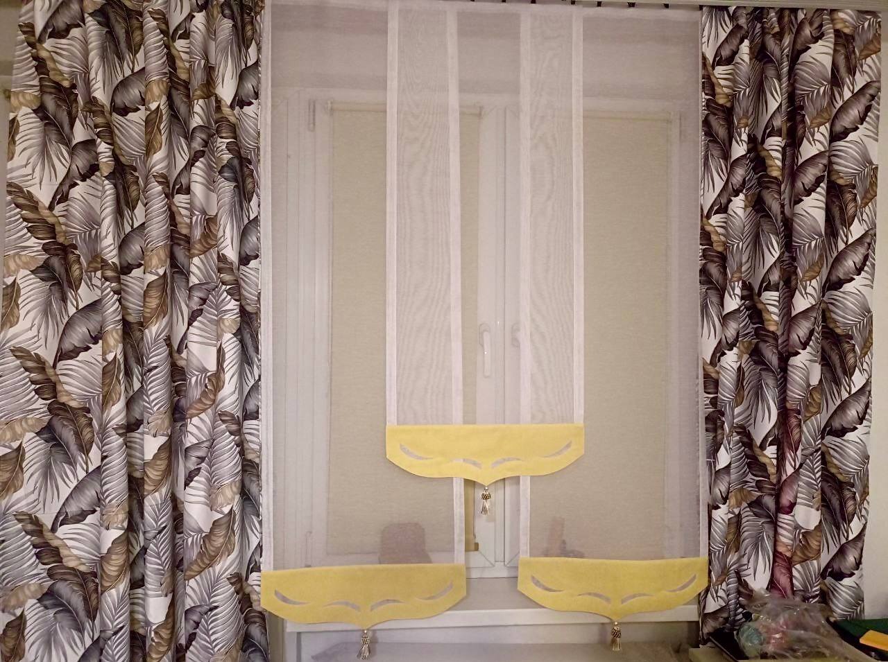 Okno z białymi, transparentnymi zasłonami z żółtymi falbanami i zasłonami po bokach w motywie liści palmowych.