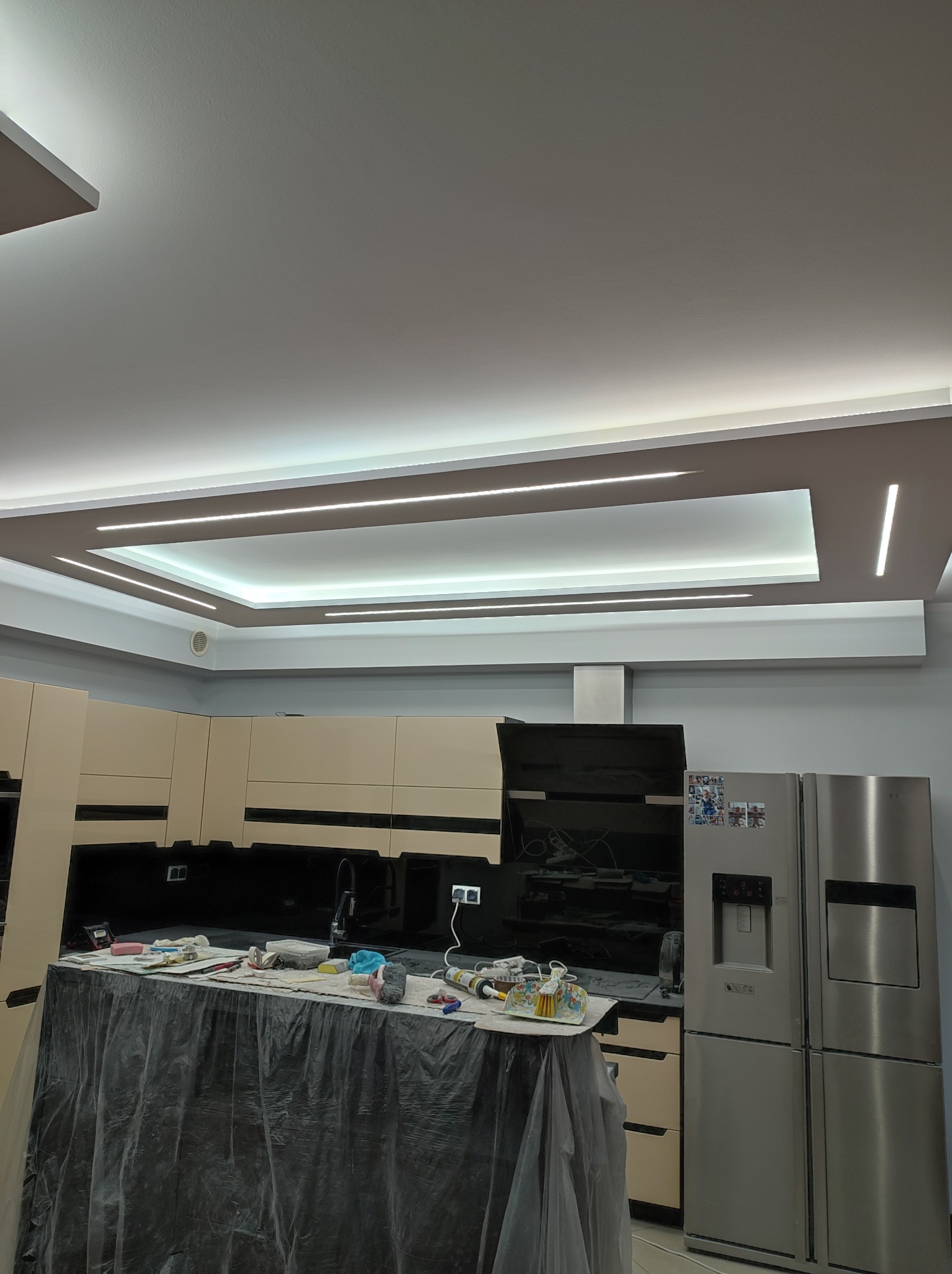 Remontowana kuchnia z osłoniętym blatem, widoczne narzędzia i akcesoria malarskie, nowoczesne oświetlenie sufitowe, zabudowa kuchenna w jasnych kolorach i srebrna lodówka.