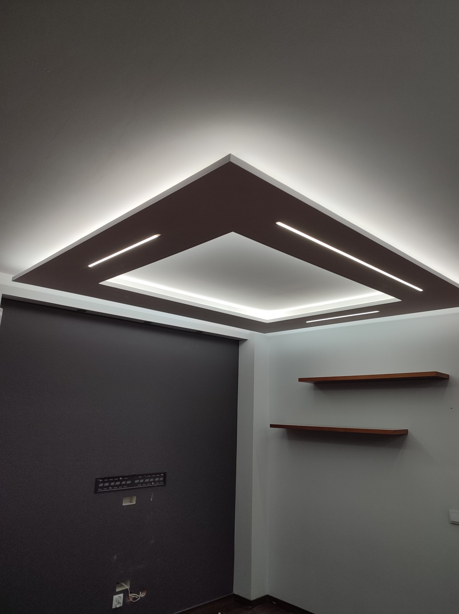 Kraków: Nowoczesny sufit podwieszany z oświetleniem LED w kształcie kwadratu, brązowe elementy dekoracyjne, minimalistyczne półki na ścianie, czarna ściana w tle.