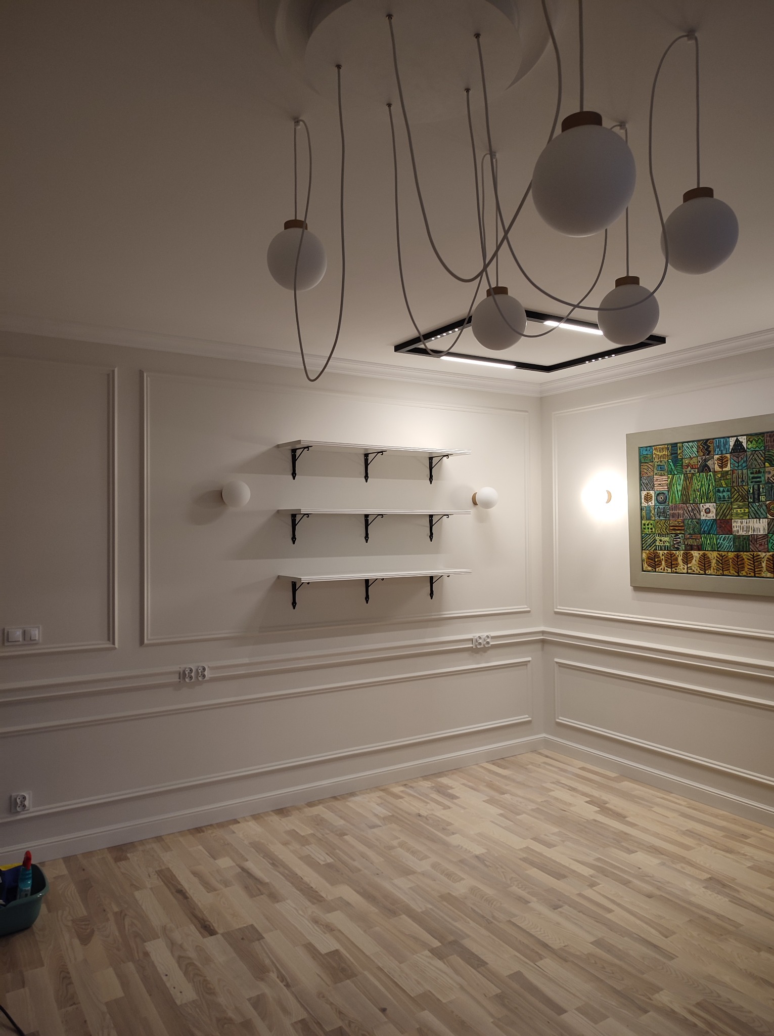 Puste, jasne pomieszczenie z drewnianą podłogą, sztukaterią na ścianach, trzema białymi półkami z czarnymi wspornikami, nowoczesnym oświetleniem sufitowym z okrągłymi kloszami i kwadratową lampą...