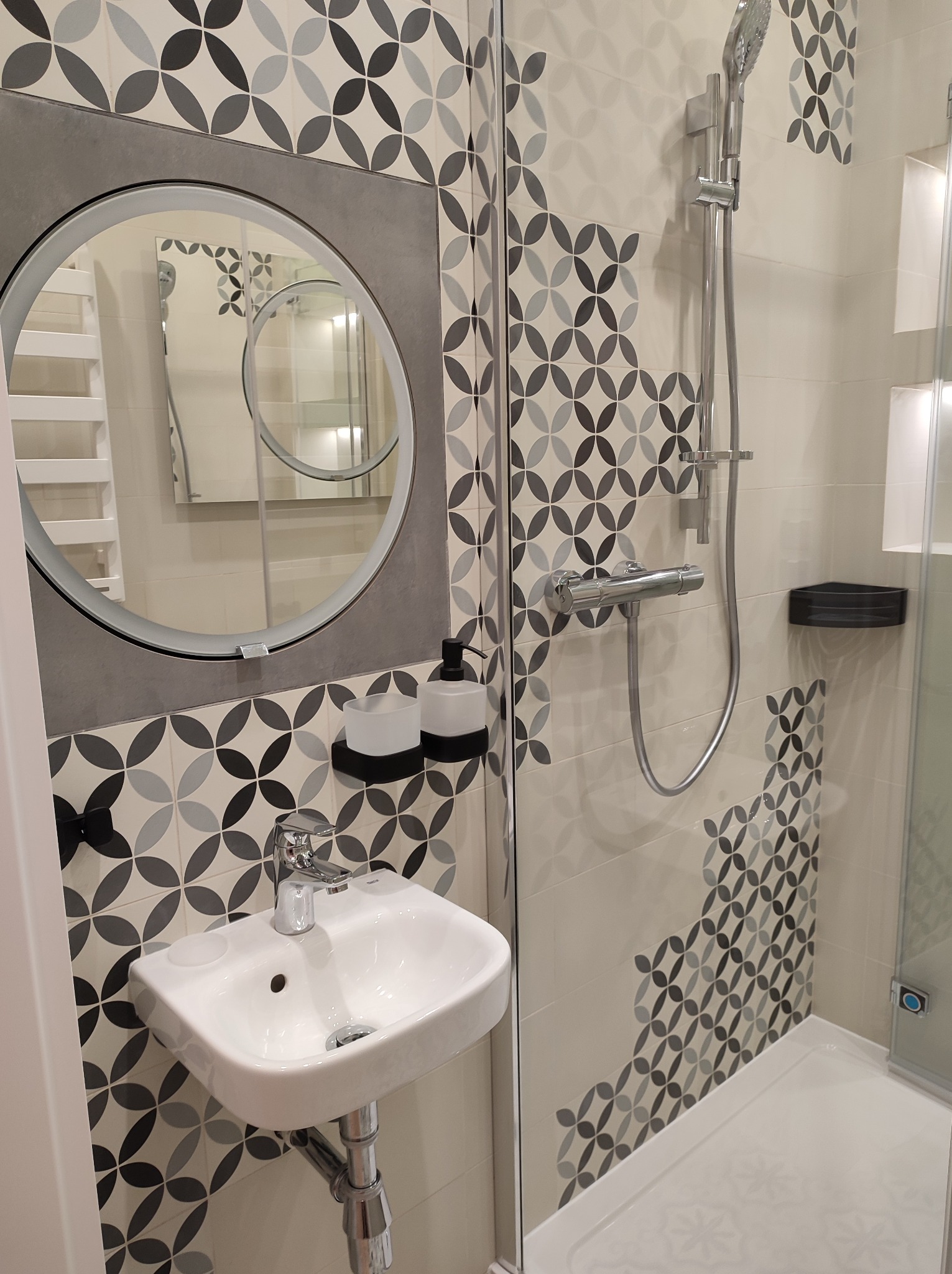 Nowoczesna łazienka z okrągłym lustrem, umywalką nablatową, kabiną prysznicową i płytkami w geometryczny wzór.