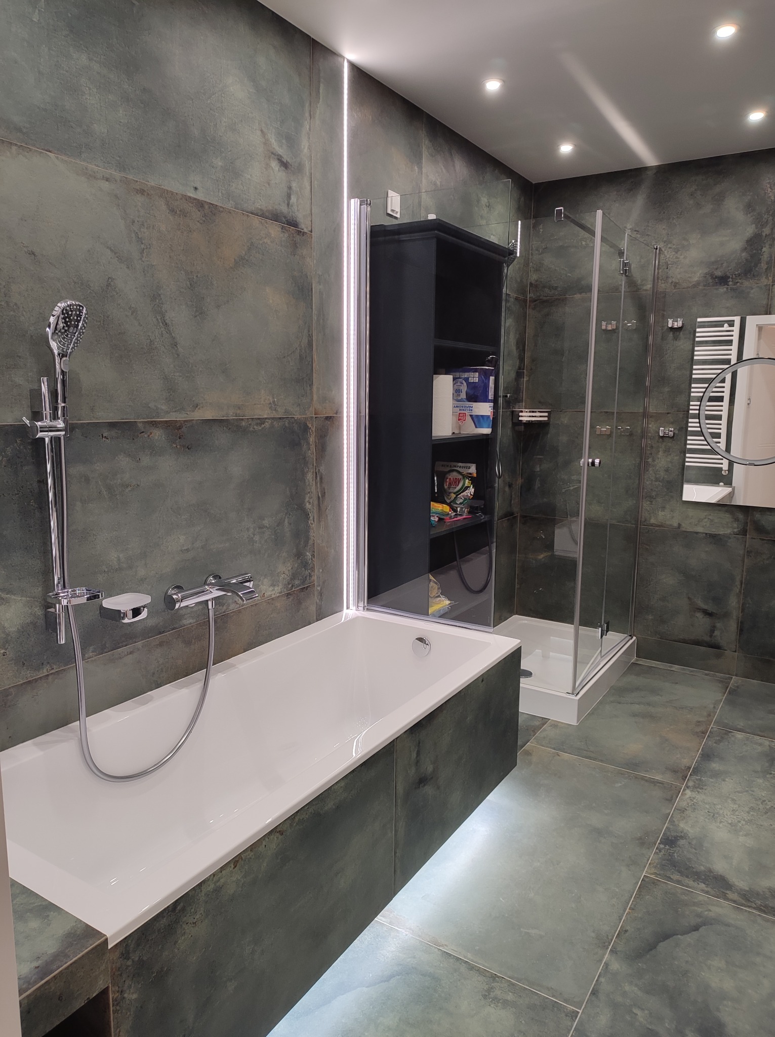 Nowoczesna łazienka z wanną, kabiną prysznicową i ciemnozielonymi płytkami imitującymi kamień, podświetlenie LED pod wanną, szafka z półkami na akcesoria łazienkowe.