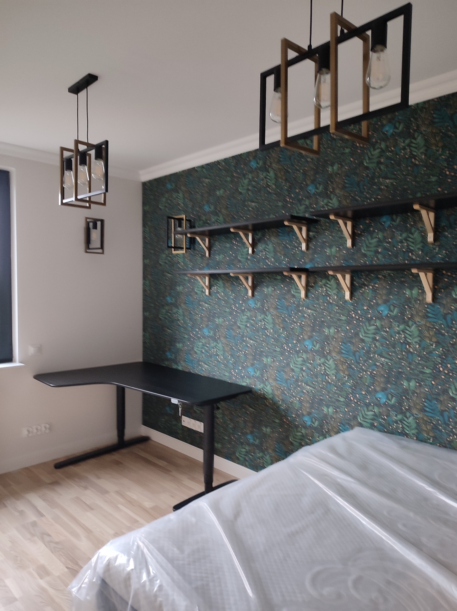 Nowoczesny pokój z czarnym biurkiem z regulowaną wysokością, półkami na tle wzorzystej tapety i designerskimi lampami wiszącymi; meble częściowo zakryte folią ochronną.