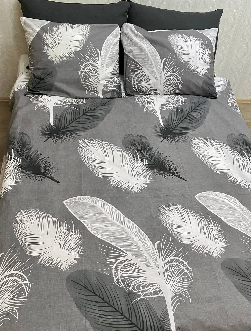 Rozłożona pościel w odcieniach szarości z motywem piór, dwa czarne poduszki w tle.