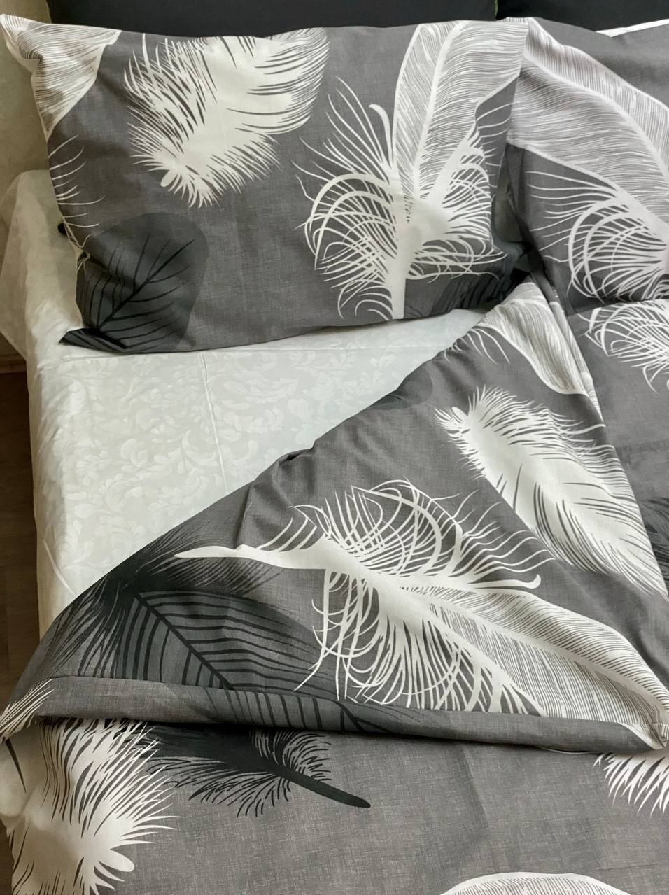 Komplet pościeli w odcieniach szarości z motywem piór, widoczna poduszka i kołdra na białym prześcieradle z delikatnym wzorem.