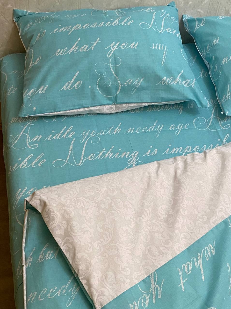 Turkusowa pościel z białym napisem i wzorzystą białą kołdrą, widoczne poduszki w tym samym stylu.