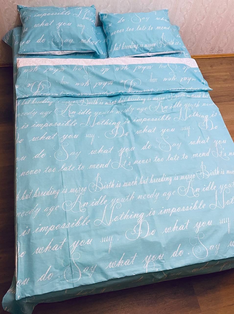 Komplet pościeli w odcieniach błękitu z białym napisem kaligraficznym, ułożony na drewnianej podłodze, składający się z dwóch poduszek i kołdry z widocznym białym prześcieradłem.