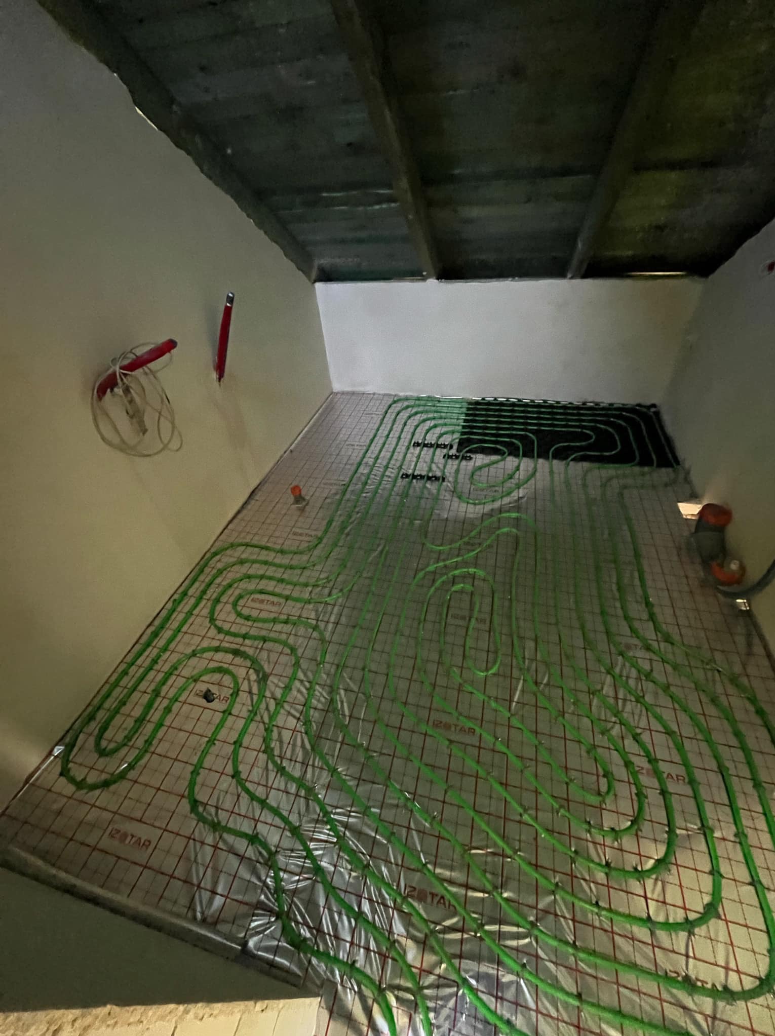 Instalacja ogrzewania podłogowego z zielonych rurek na folii aluminiowej w surowym pomieszczeniu z betonowym stropem i widocznymi przewodami elektrycznymi na ścianie.