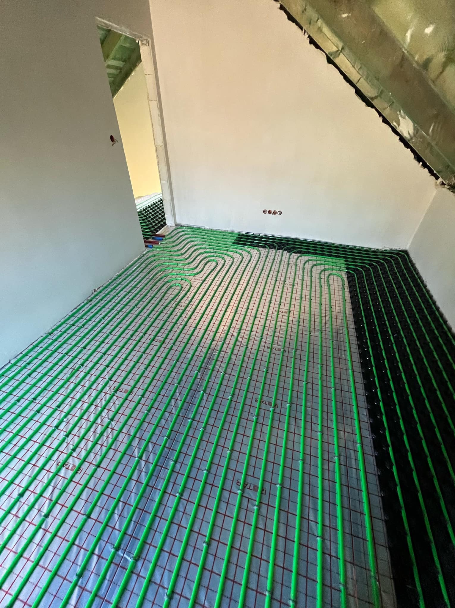 Wnętrze w trakcie instalacji ogrzewania podłogowego: zielone rurki ułożone na srebrnej folii odblaskowej, widoczne czarne rurki przy ścianie, surowy stan wykończenia ścian i poddasza.