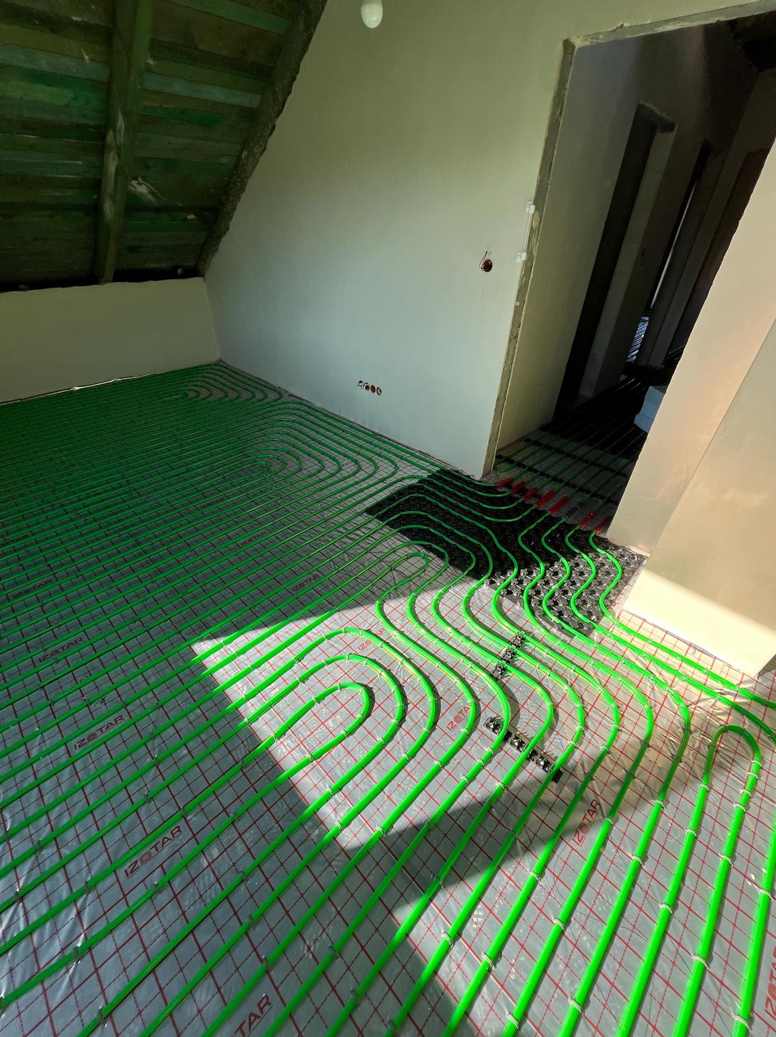 Wnętrze w trakcie montażu ogrzewania podłogowego: zielone rurki grzewcze ułożone na folii z siatką w pomieszczeniu ze skośnym sufitem i widoczną instalacją elektryczną.
