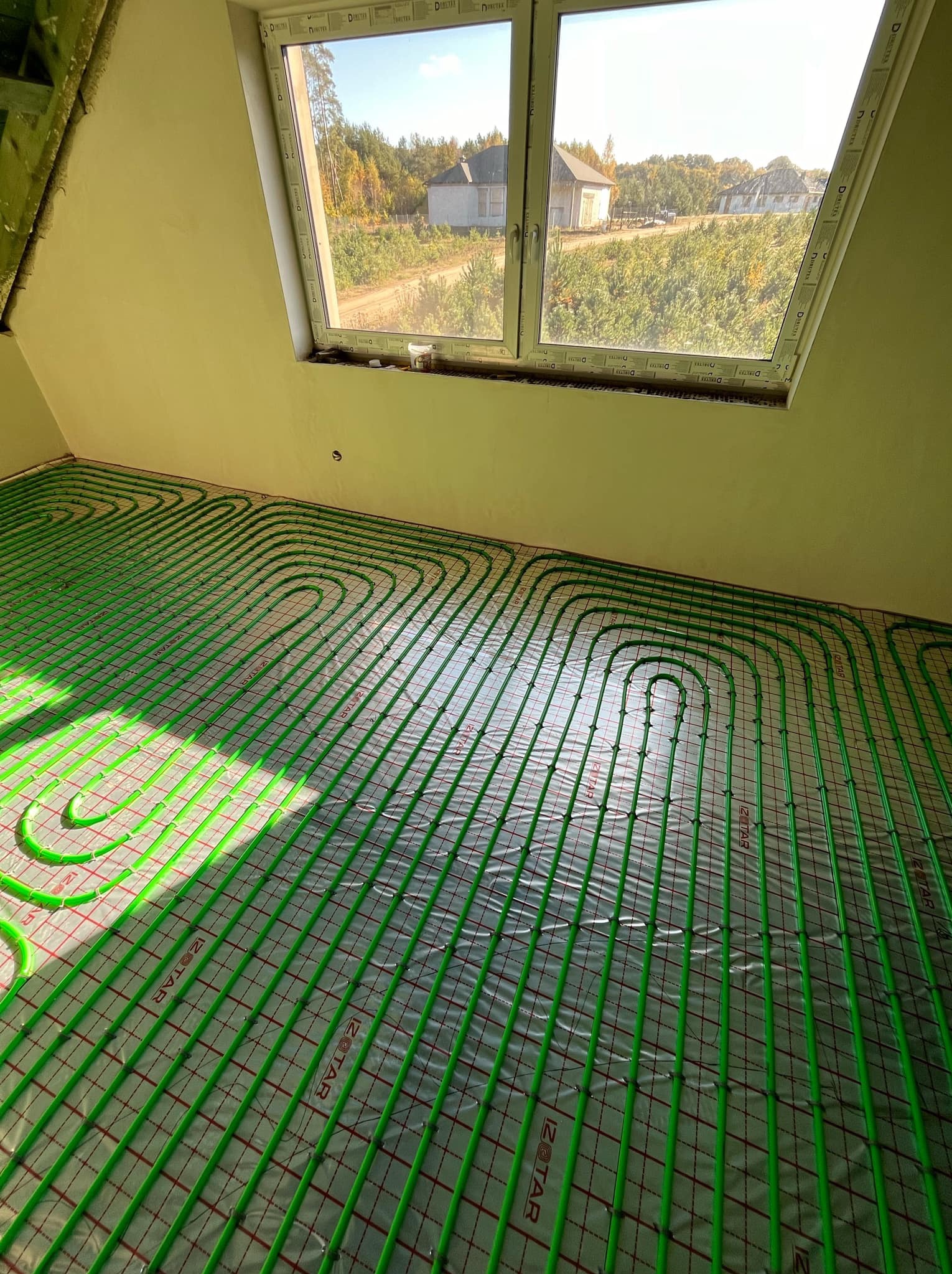 Zielone rury ogrzewania podłogowego ułożone na folii z siatką w jasnym pokoju z oknem dachowym, widok na wiejski krajobraz