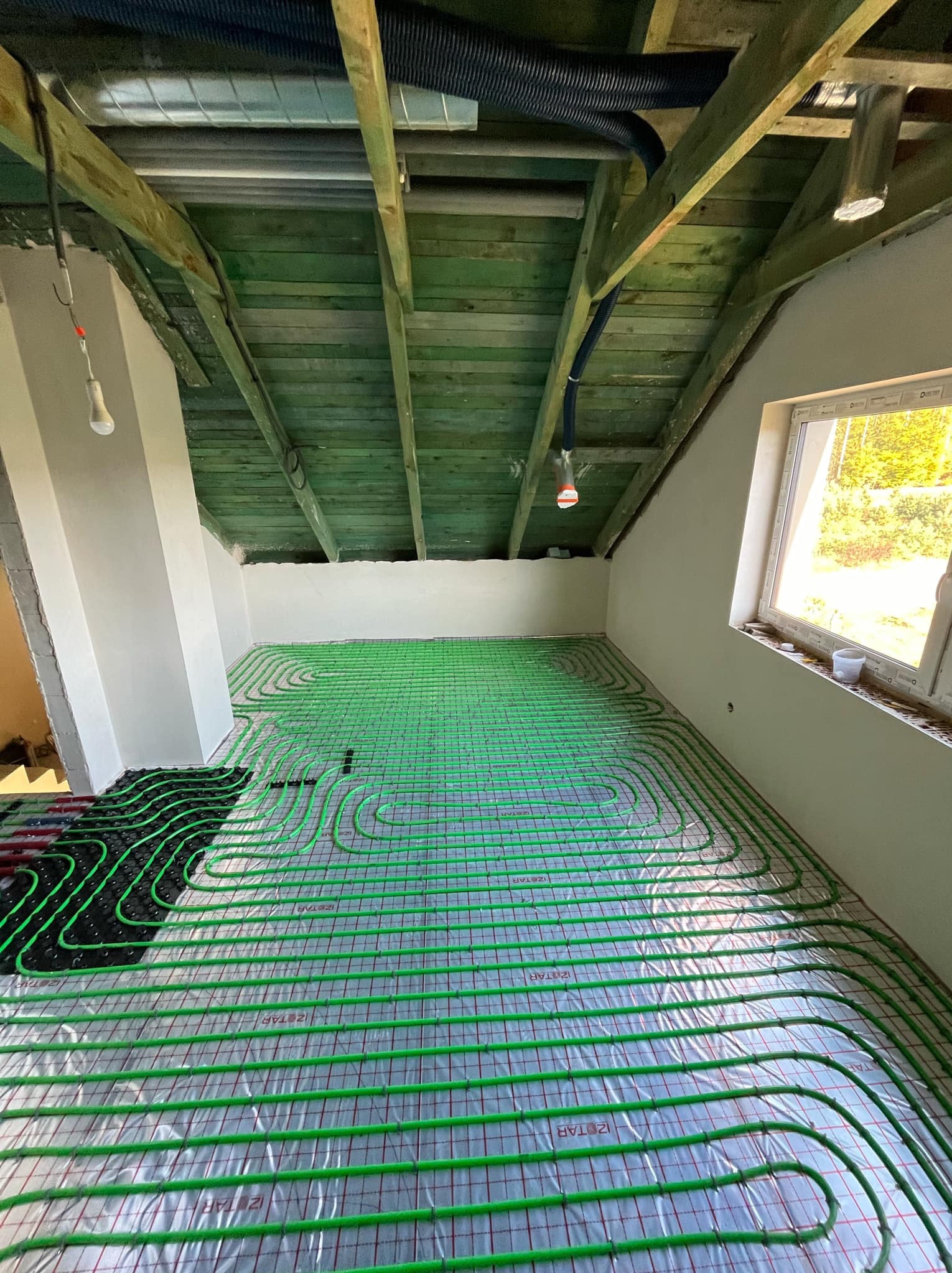 Zielone rury ogrzewania podłogowego ułożone na folii w pomieszczeniu z surowym drewnianym stropem i oknem z widokiem na zieleń.