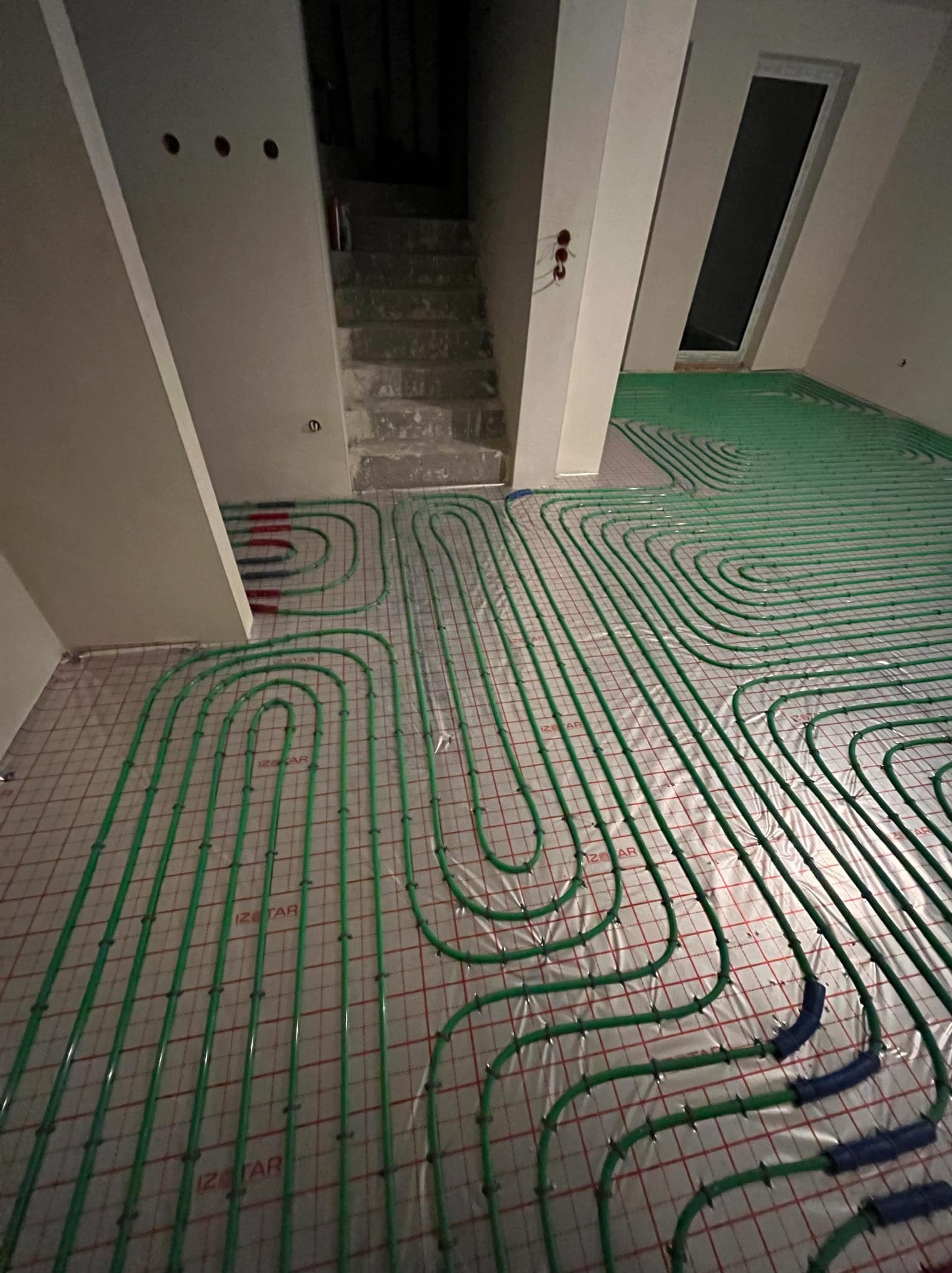 Zielone rury ogrzewania podłogowego układane na folii z siatką w nowym budynku, widoczne schody w tle.