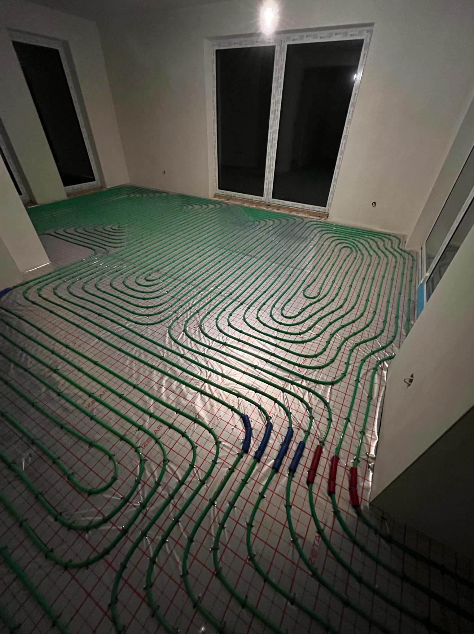 Zielone rurki ogrzewania podłogowego ułożone na srebrnej folii z czerwoną siatką w jasnym pomieszczeniu z dużymi oknami.