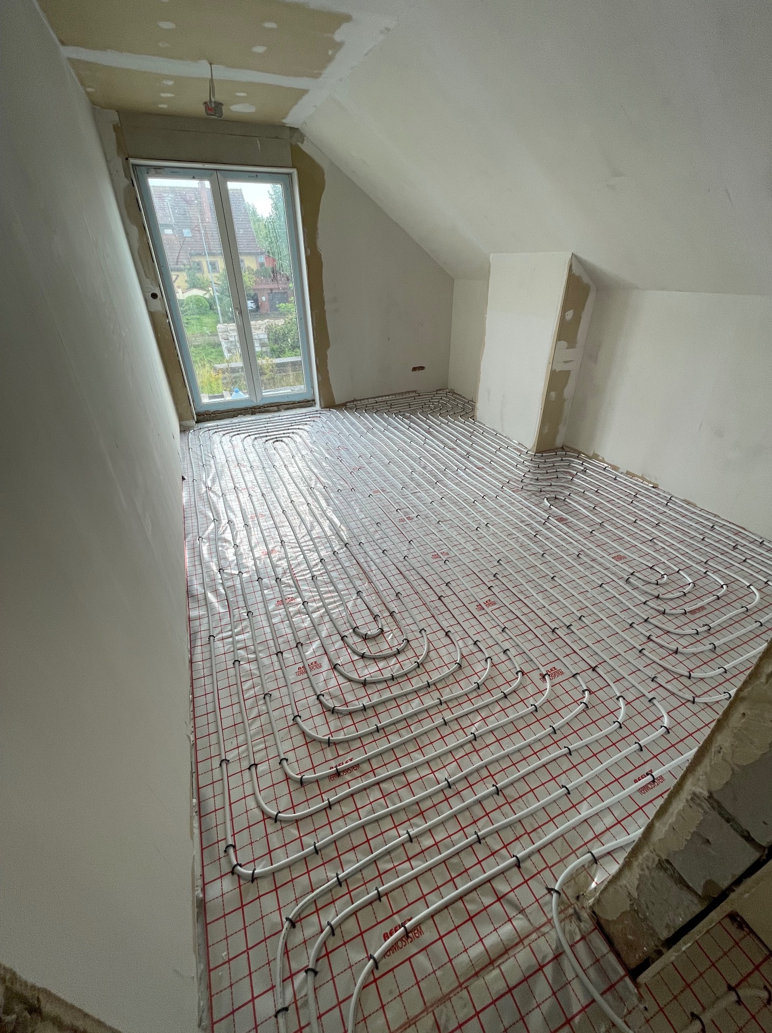 Instalacja ogrzewania podłogowego w nowym domu na poddaszu, rury grzewcze ułożone na folii z czerwoną siatką, widok na okno i surowy stan ścian.