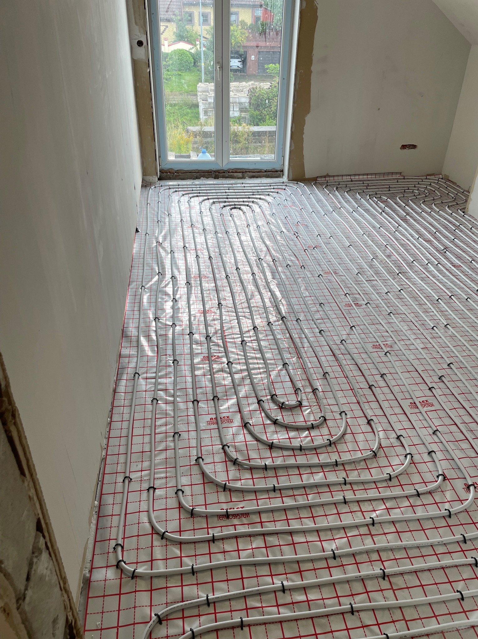 Białe rury ogrzewania podłogowego ułożone na folii z czerwoną kratką w surowym stanie deweloperskim, widok z góry na instalację przed wylewką.