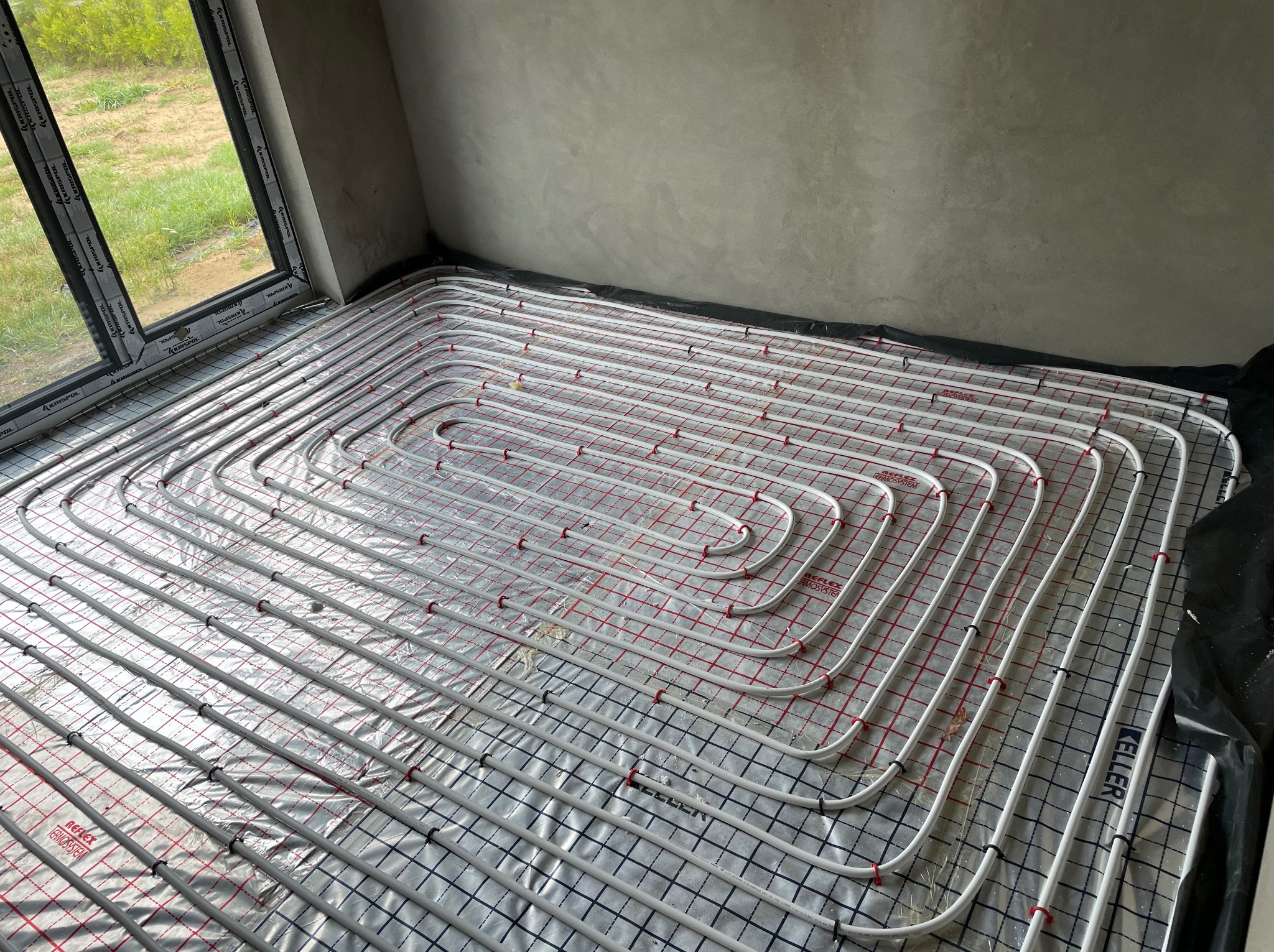 Układanie białych rur ogrzewania podłogowego na folii aluminiowej z czerwonymi i niebieskimi liniami siatki w surowym wnętrzu z oknem.