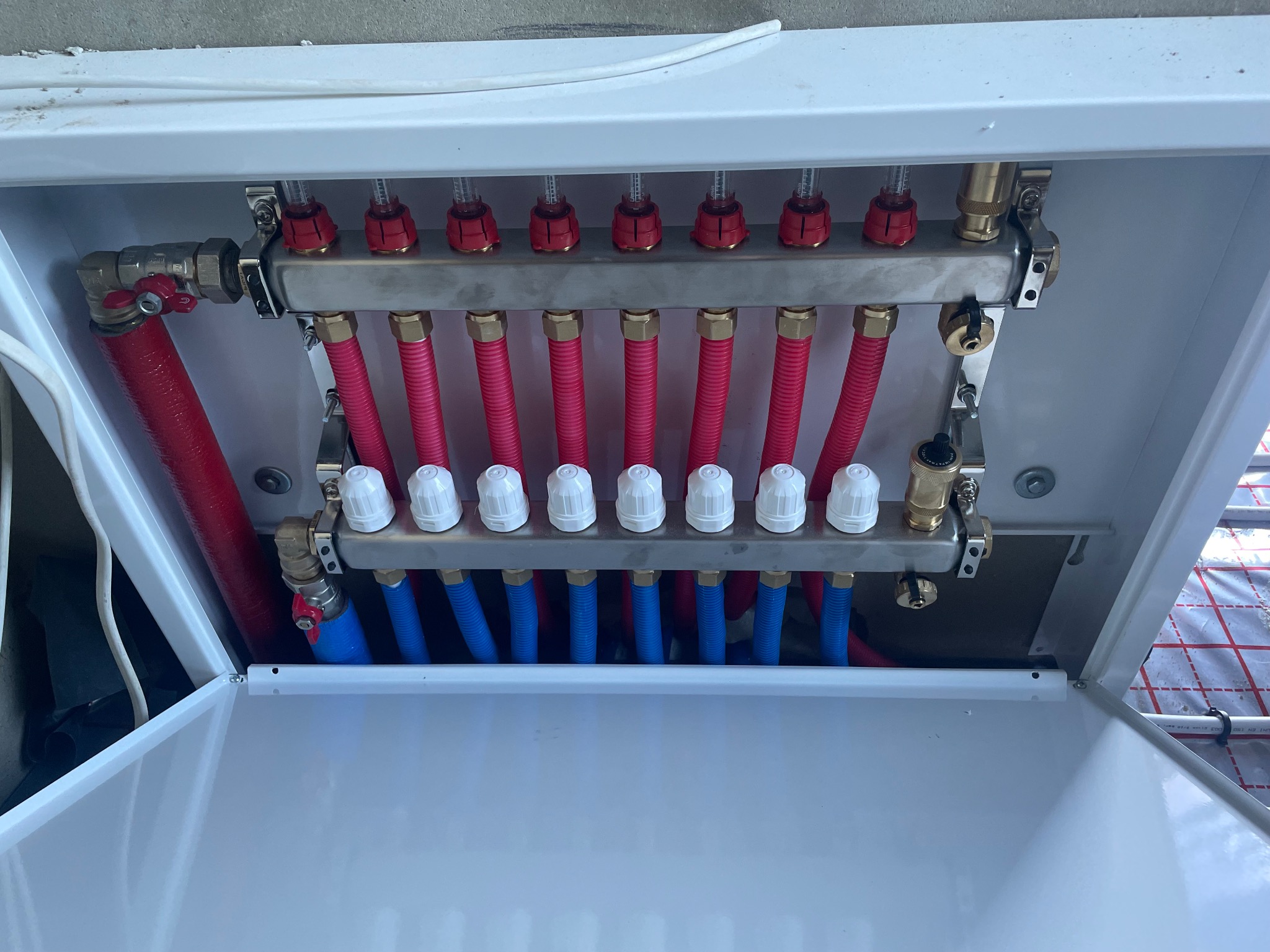 Rozdzielacz ogrzewania podłogowego w białej szafce, widoczne czerwone i niebieskie rury oraz regulatory przepływu.