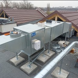 PROWENT INSTALL DENYS SHOLOMYTSKYI - Montaż rekuperacji na dachu budynku komercyjnego: widoczny pracownik z narzędziami, system rekuperacji z metalowymi rurami i elementami mocującymi na betonowych podstawach, w tle dachy budynków...