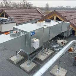 Montaż rekuperacji na dachu budynku komercyjnego: widoczny pracownik z narzędziami, system rekuperacji z metalowymi rurami i elementami mocującymi na betonowych podstawach, w tle dachy budynków...