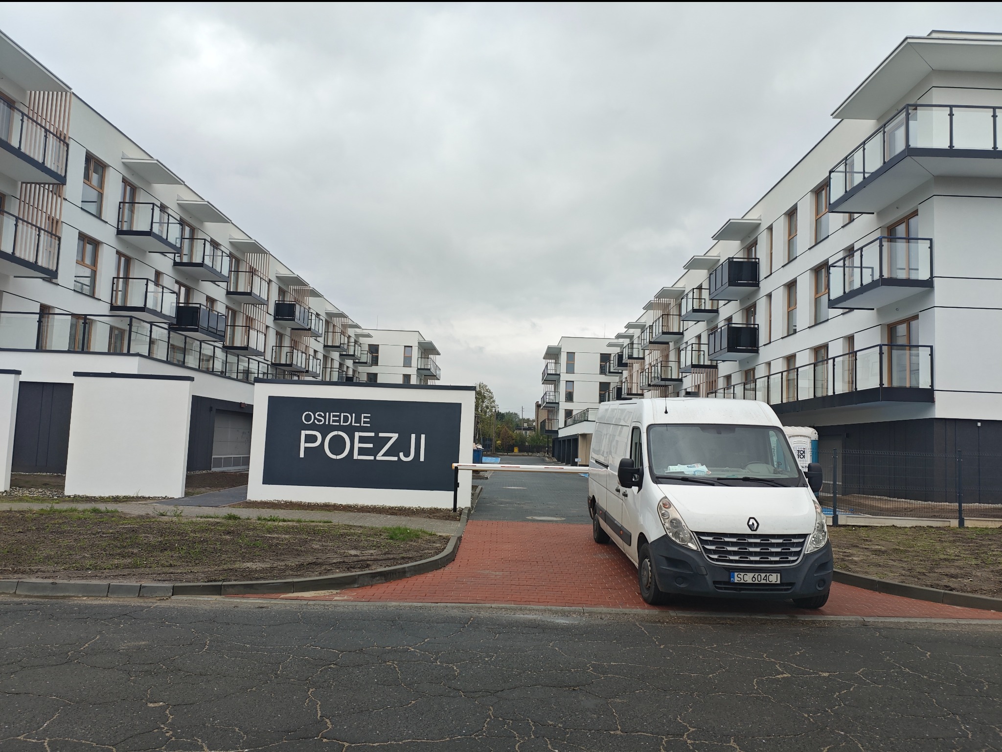 Nowoczesne budynki apartamentowe z balkonami, brama wjazdowa z napisem 'Osiedle Poezji' i zaparkowany biały van na tle pochmurnego nieba.