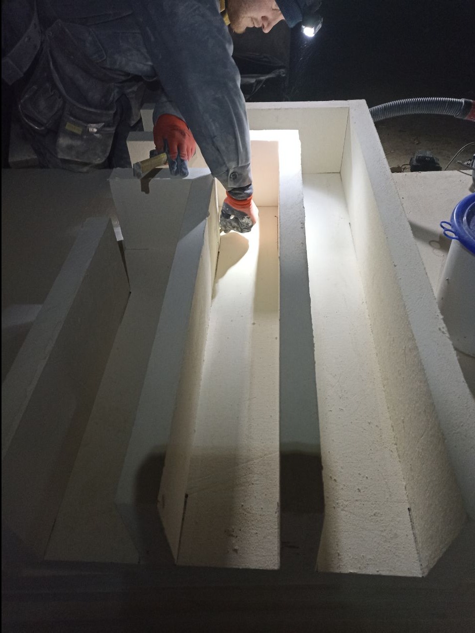 Pracownik w kombinezonie i rękawicach montuje białe elementy izolacyjne w systemie kanałów wentylacyjnych, oświetlony lampą czołową.