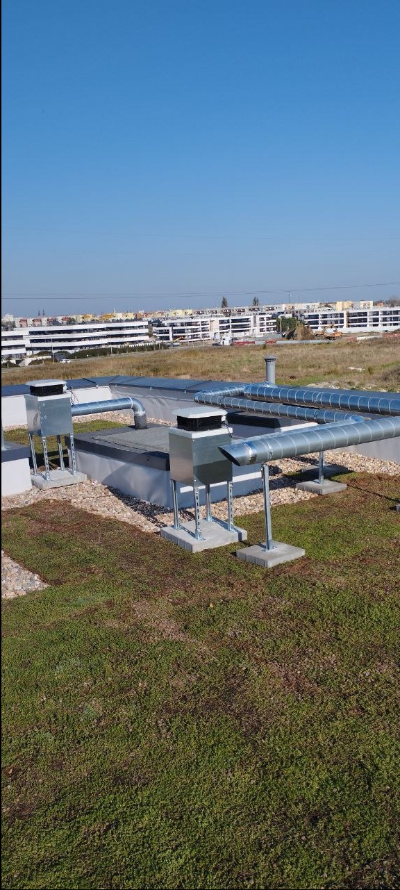 Instalacja wentylacyjna z metalowymi rurami na zielonym dachu budynku komercyjnego, widok na miasto w tle.