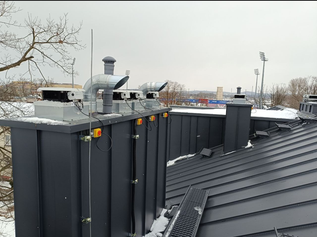 Kompletna instalacja wentylacyjna z rekuperacją na dachu budynku, widoczne przewody, anemostaty i elementy sterujące, w tle zabudowa miejska i stadion.