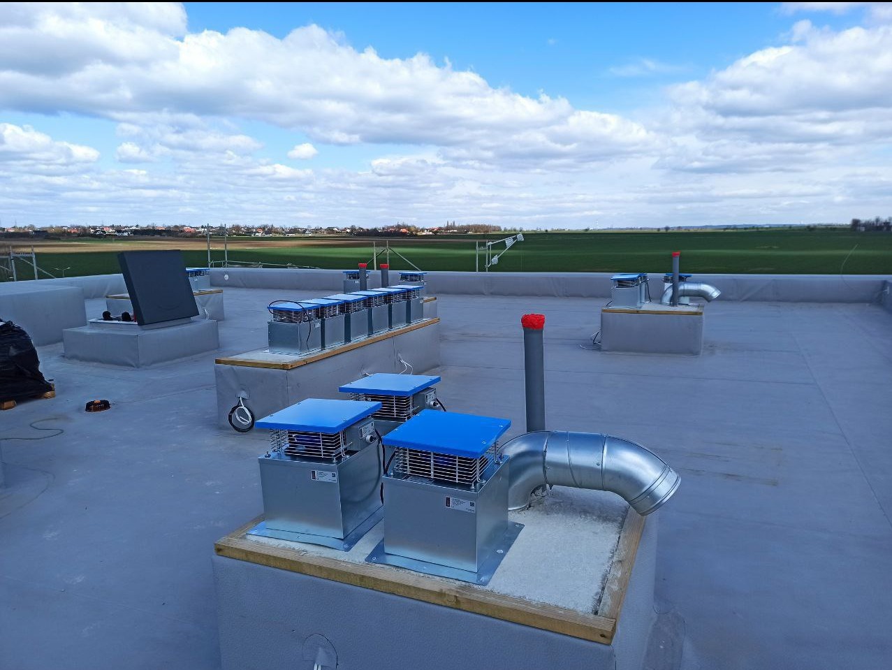 Widok z góry na instalację wentylacyjną na dachu budynku komercyjnego, z licznymi srebrnymi jednostkami wentylacyjnymi z niebieskimi pokrywami i rurami, na tle zielonych pól i błękitnego nieba...