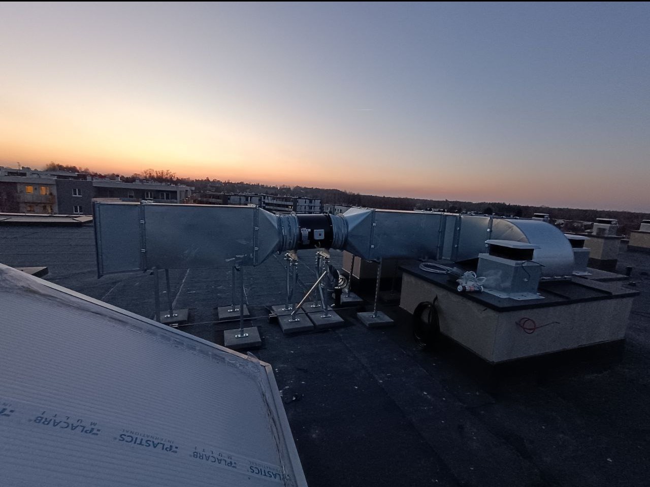 System kanałów wentylacyjnych na dachu budynku komercyjnego o zmierzchu, widoczne elementy montażowe i konstrukcja wsporcza.