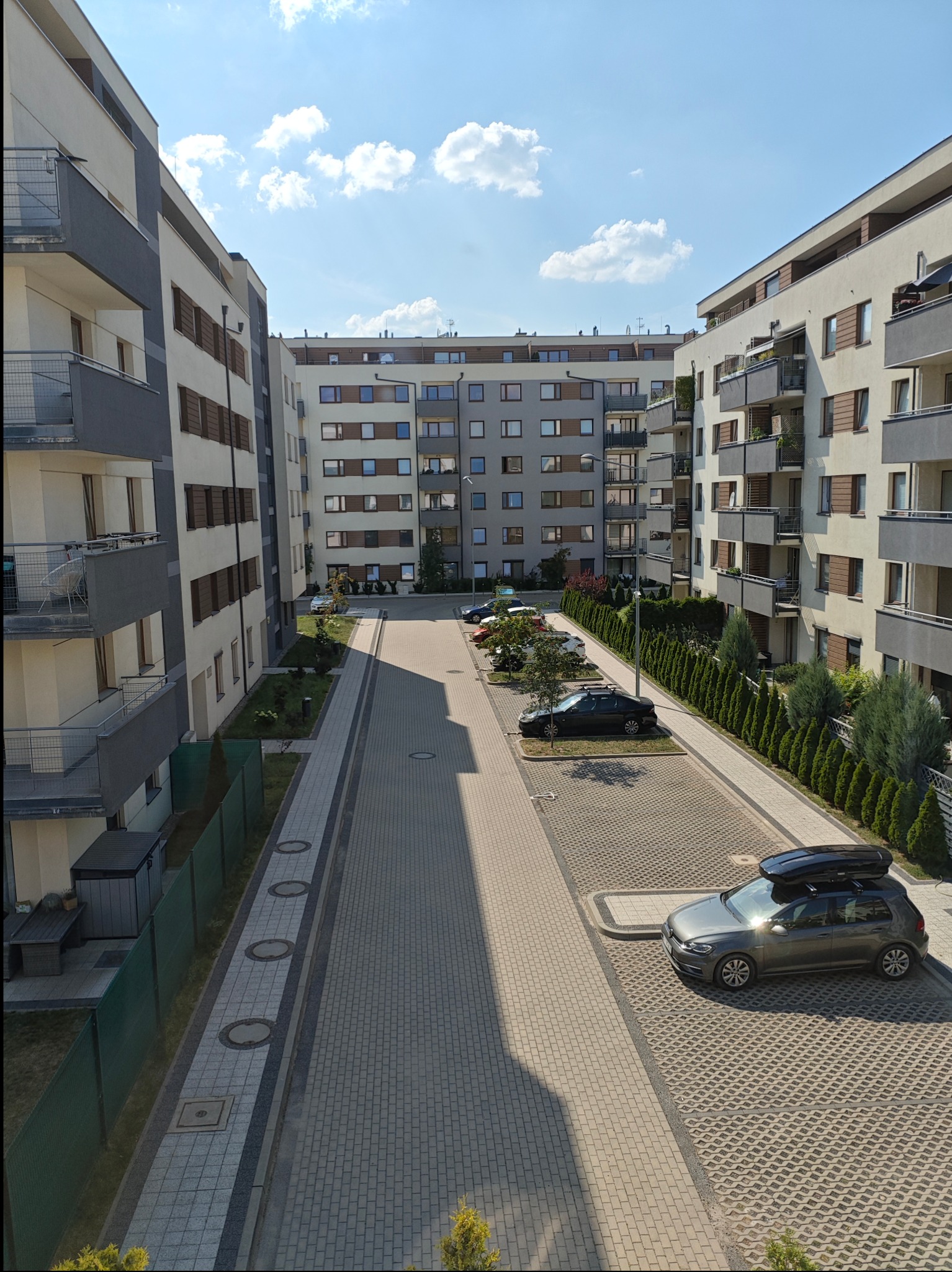 Widok z góry na dziedziniec osiedla mieszkaniowego z zaparkowanymi samochodami i zielenią, w tle budynki z balkonami, jasne niebo z chmurami.