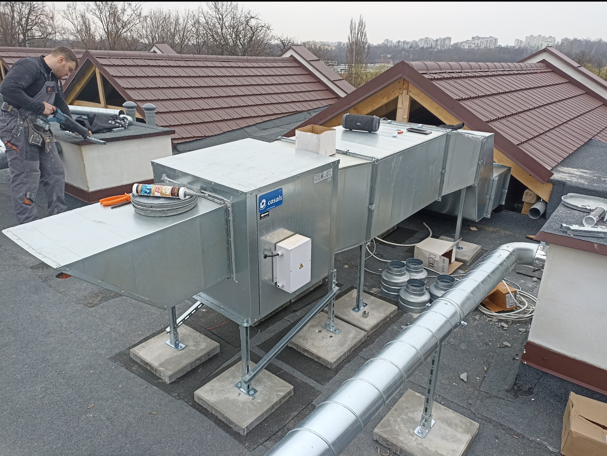 Montaż rekuperacji na dachu budynku komercyjnego: widoczny pracownik z narzędziami, system rekuperacji z metalowymi rurami i elementami mocującymi na betonowych podstawach, w tle dachy budynków...