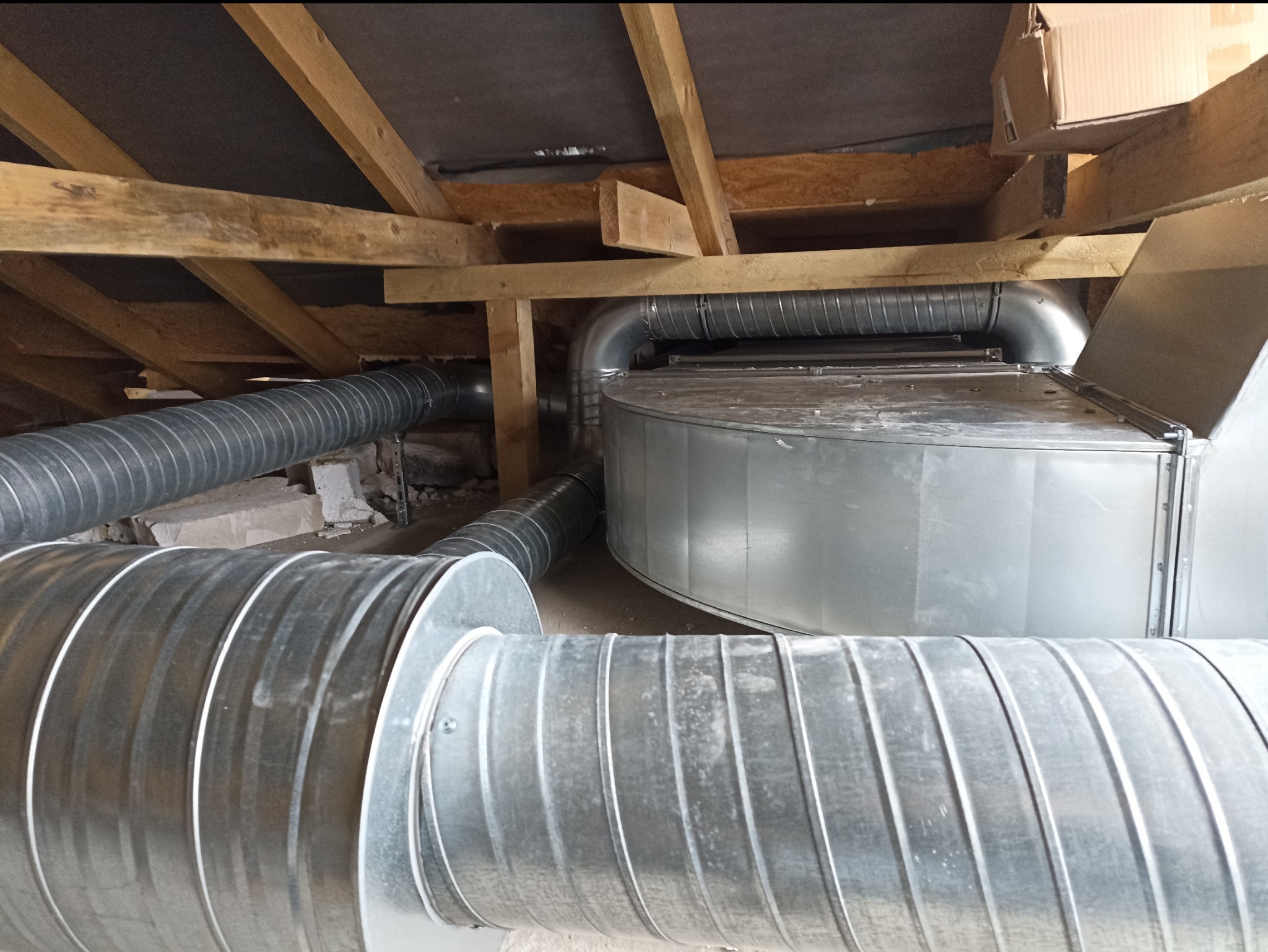 Szare, metalowe rury wentylacyjne karbowane, zamontowane pod drewnianą konstrukcją dachu na poddaszu, widoczne fragmenty muru i karton.