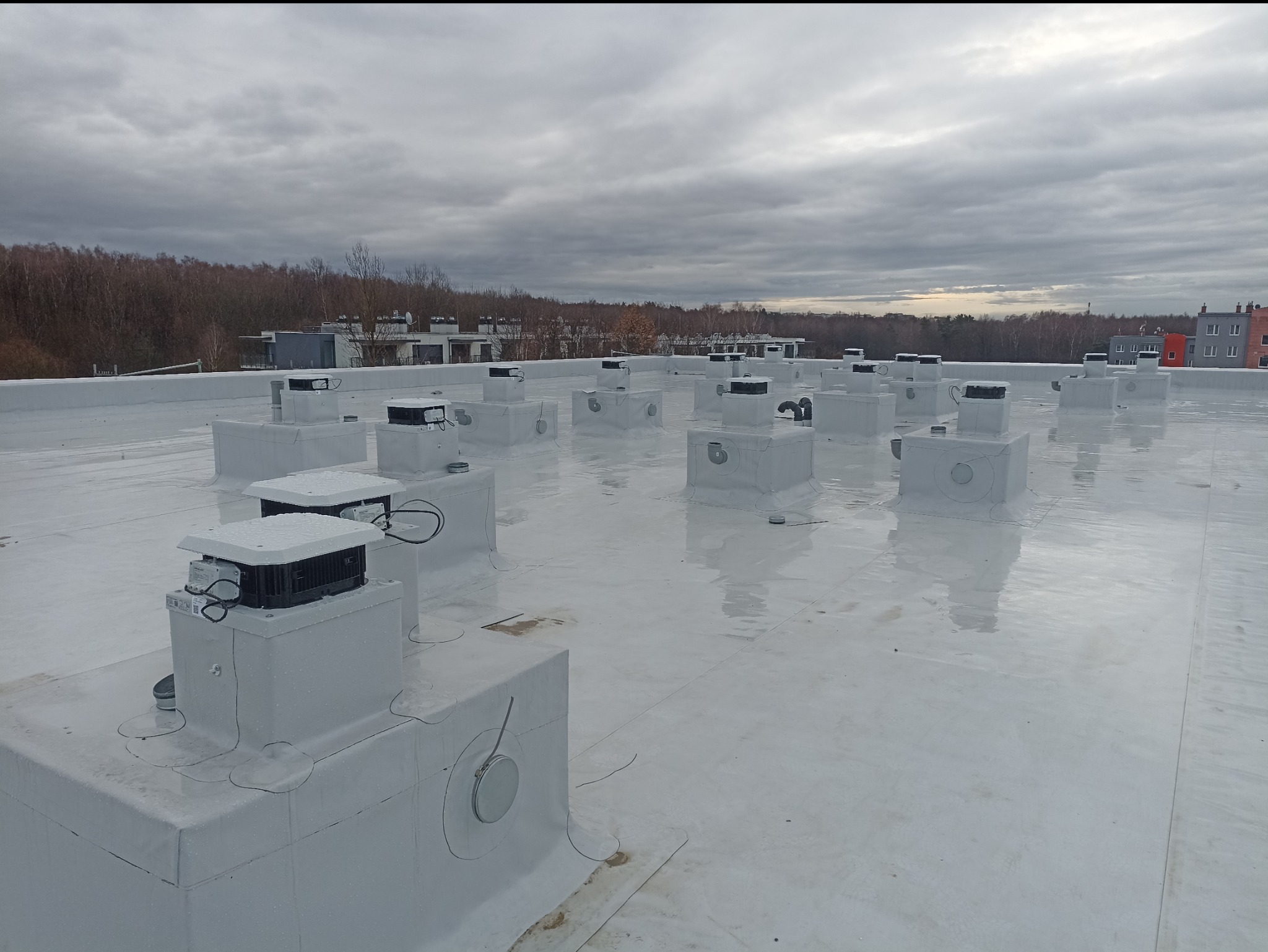 Szereg białych, kwadratowych obudów wentylacyjnych z czarnymi elementami na płaskim, białym dachu budynku komercyjnego, widoczne przewody i elementy mocujące, pochmurne niebo w tle.