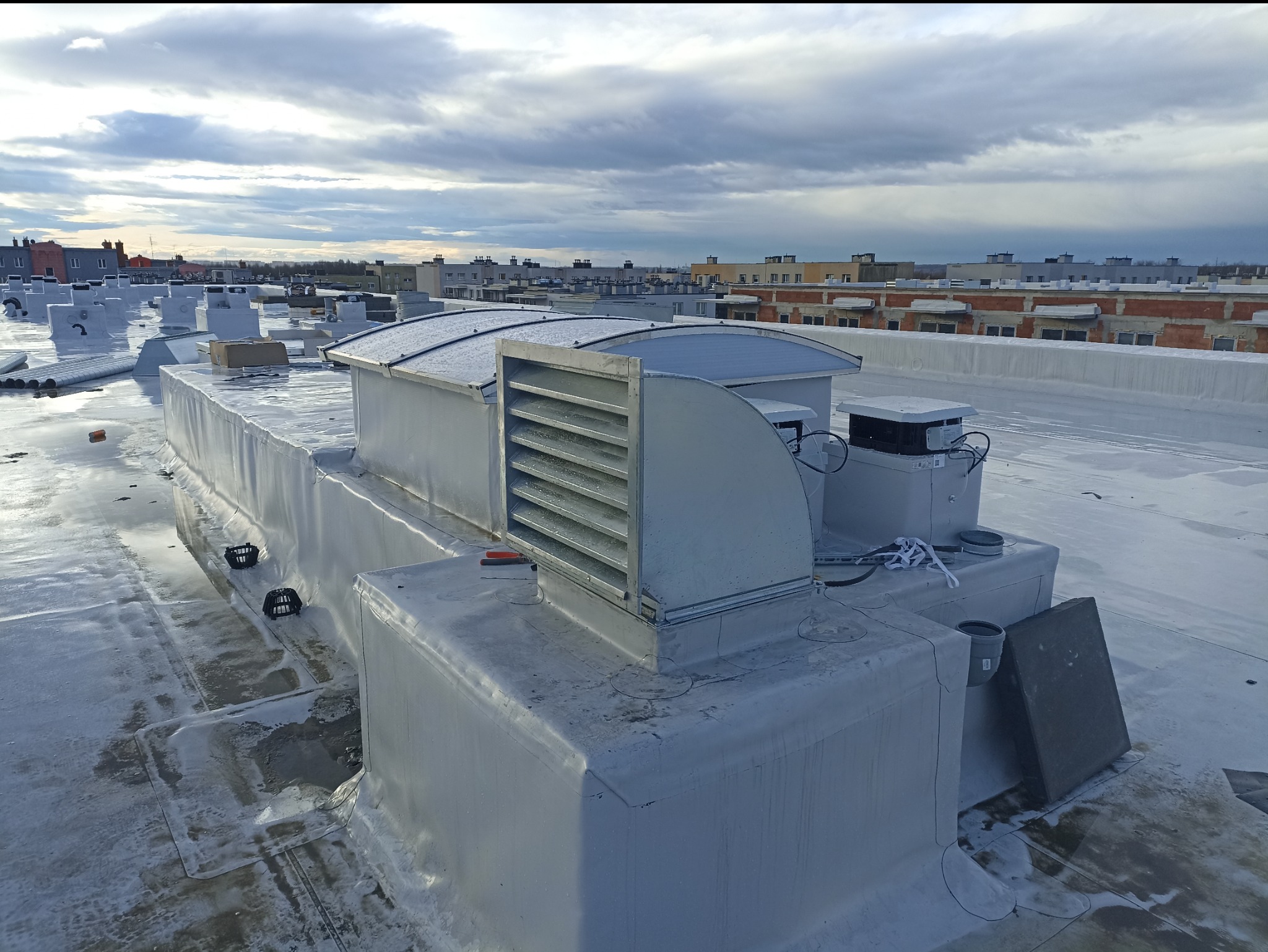Instalacja wentylacyjna na płaskim dachu budynku komercyjnego, widoczne elementy rekuperacji, kratki wentylacyjne, rury i obudowy, w tle panorama miasta pod pochmurnym niebem.